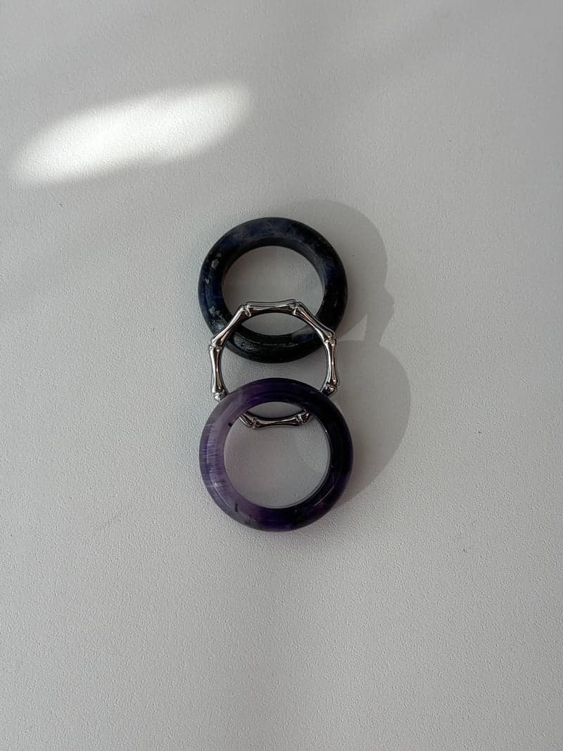 Zwei Steinringe und ein dünner silberfarbener Ring in Bambusform, senkrecht angeordnet: ein dunkelblaues Band, der silberne Ring und ein violettes Band