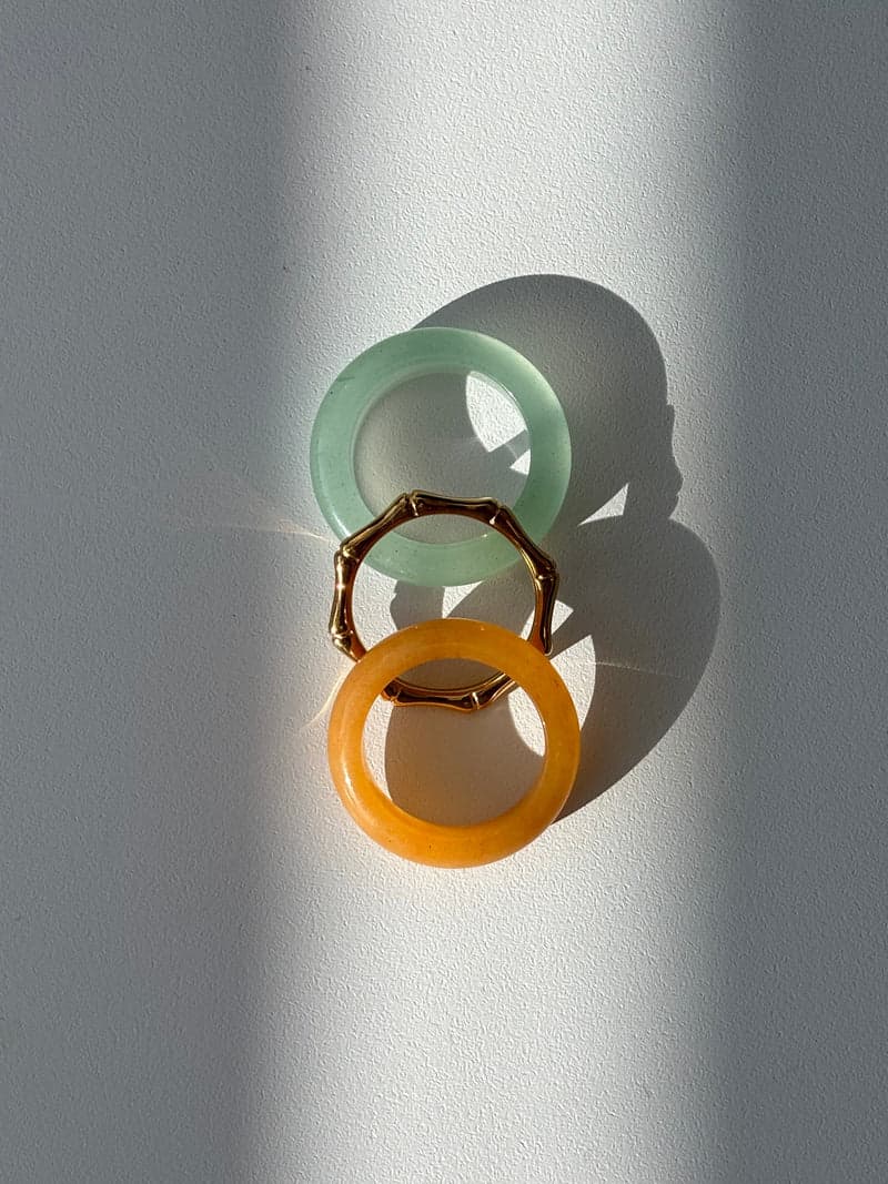 Zwei Steinbandringe, einer blassgrün und einer orange, und ein dünner goldener Ring in Bambusform, in einer Reihe angeordnet im hellen gestreiften Sonnenlicht