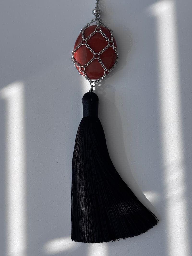 Uma pedra oval vermelho-tijolo presa em uma malha de corrente prateada acima de uma longa borla preta, contra uma parede branca sob luz listrada