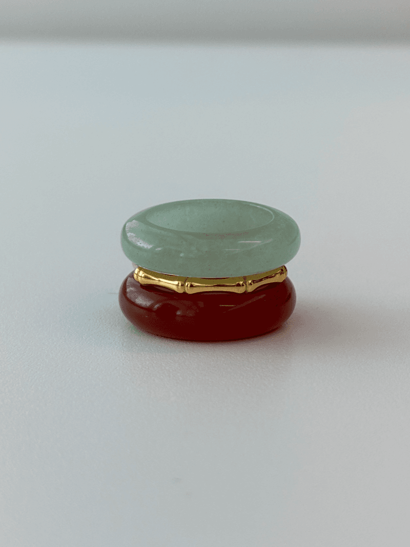 Un ensemble de bagues empilées : un anneau en pierre vert pâle au-dessus, un fin anneau doré en forme de bambou, et un anneau en pierre rouge profond en dessous
