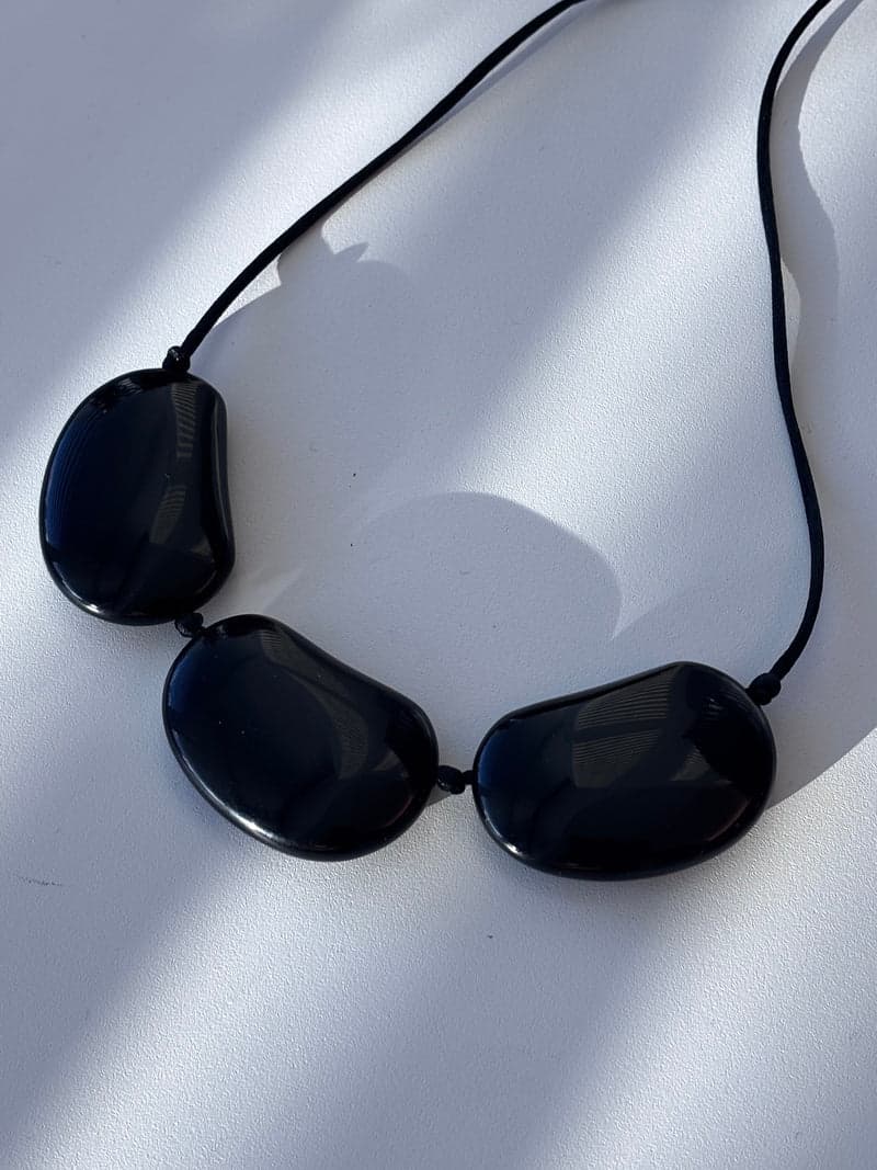 Três grandes pedras ovais achatadas, pretas e brilhantes, enfiadas em um cordão preto, sobre uma superfície branca sob luz listrada