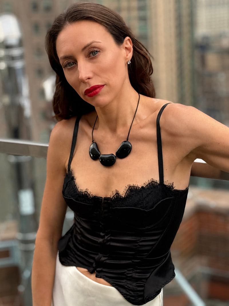 Une femme sur un balcon en ville dans un haut en dentelle noire, portant un collier de trois grandes pierres ovales sombres sur un cordon noir