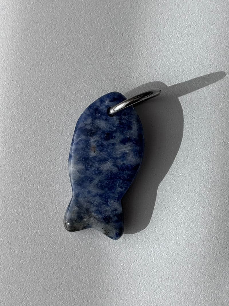 Ein kleiner geschnitzter Fisch-Anhänger aus gesprenkeltem blau-weißem Stein mit einem silbernen Ring, auf weißem Untergrund im Fensterlicht
