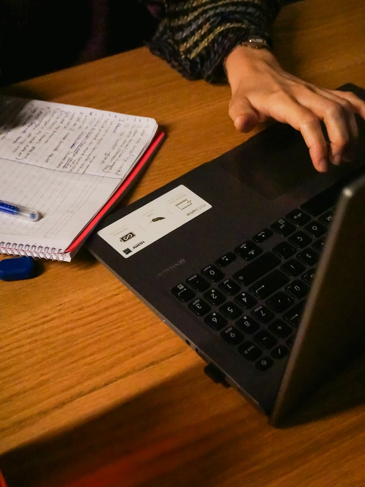 Hands typing on a laptop at a wooden desk beside an open notebook