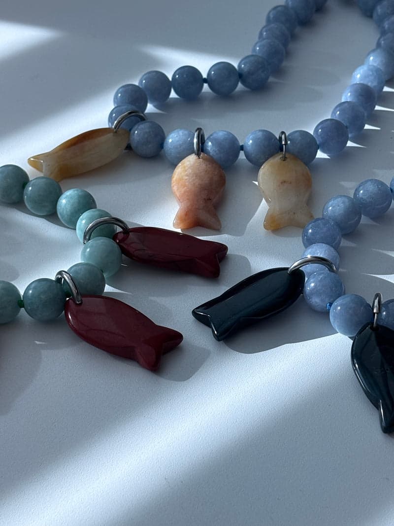 Pale green and pale blue bead necklaces laid together in window light, hung with small carved fish pendants in orange, red and dark green