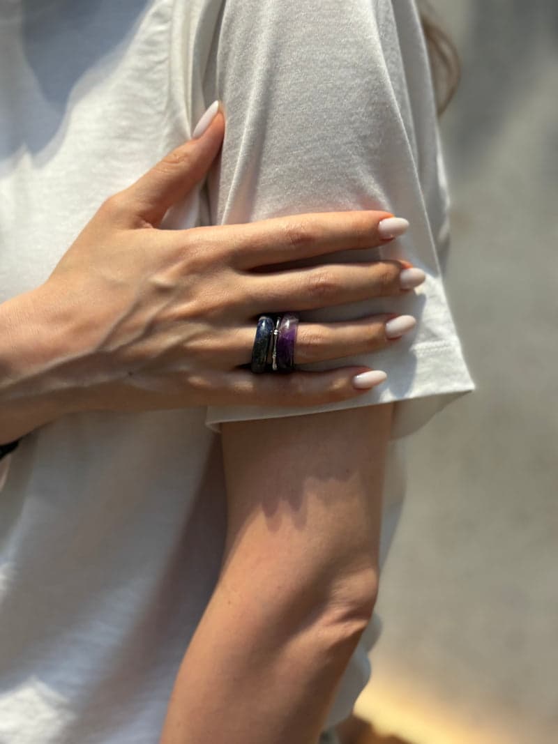Eine Hand auf einem bloßen Oberarm vor einem weißen T-Shirt, mit den violetten und dunkelblauen Steinringen, gestapelt an einem Finger