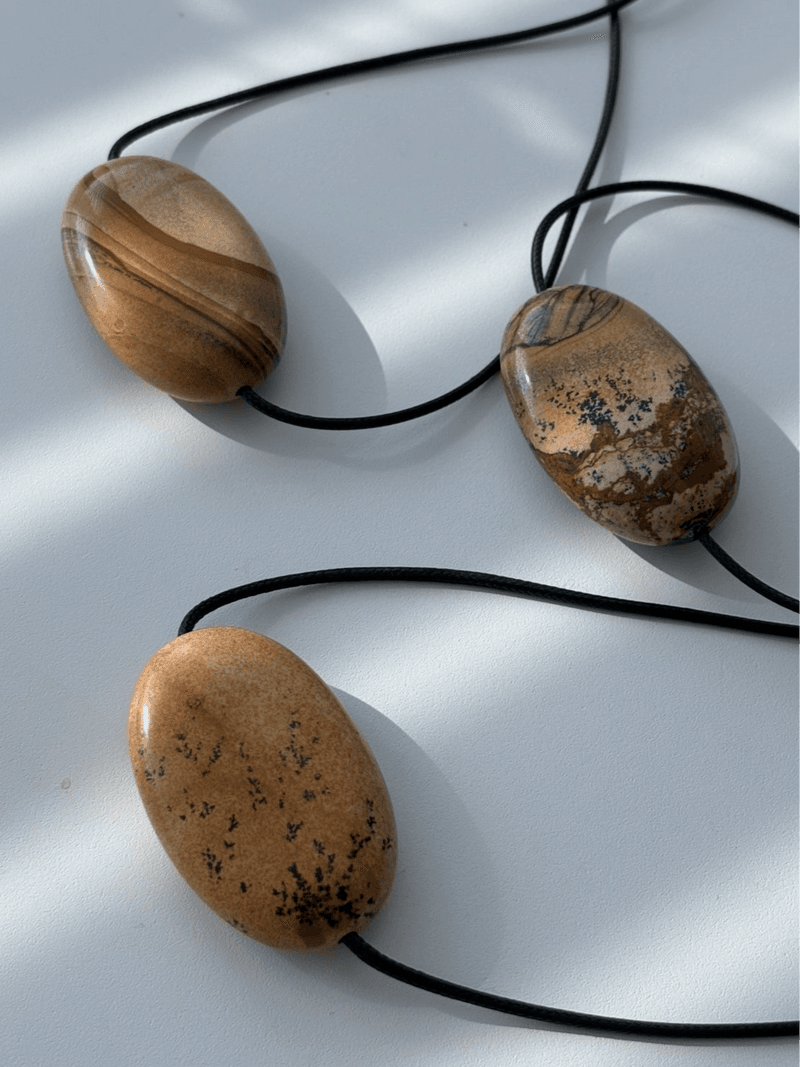 Três grandes pingentes ovais achatados em pedra bege e marrom com marcas escuras ramificadas, em cordões pretos sobre uma superfície branca