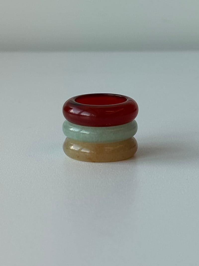 Three stone band rings stacked on a white surface: deep red on top, pale green in the middle, pale yellow below