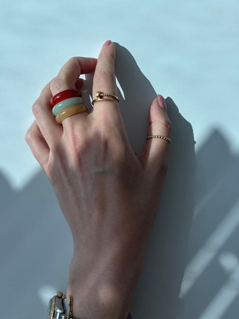 A hand against a pale blue wall wearing the red, green and yellow stone rings stacked on one finger