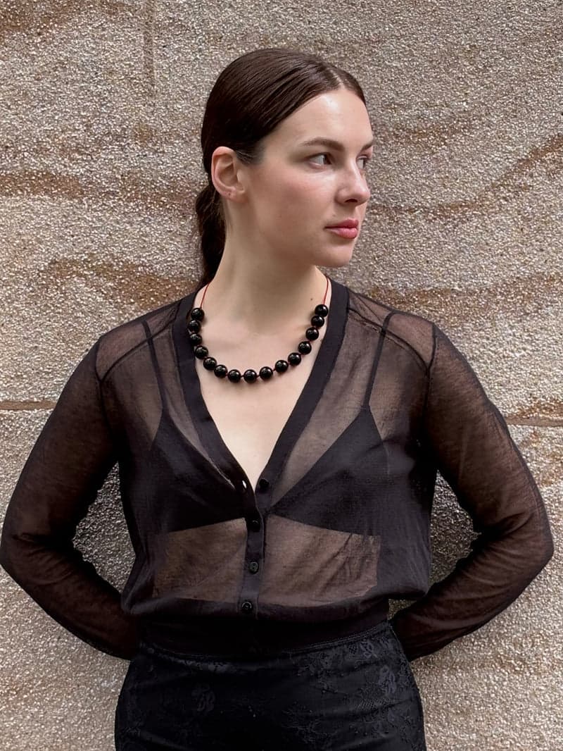 A woman in a sheer black blouse standing against a sandstone wall, wearing the black bead necklace close at the neck