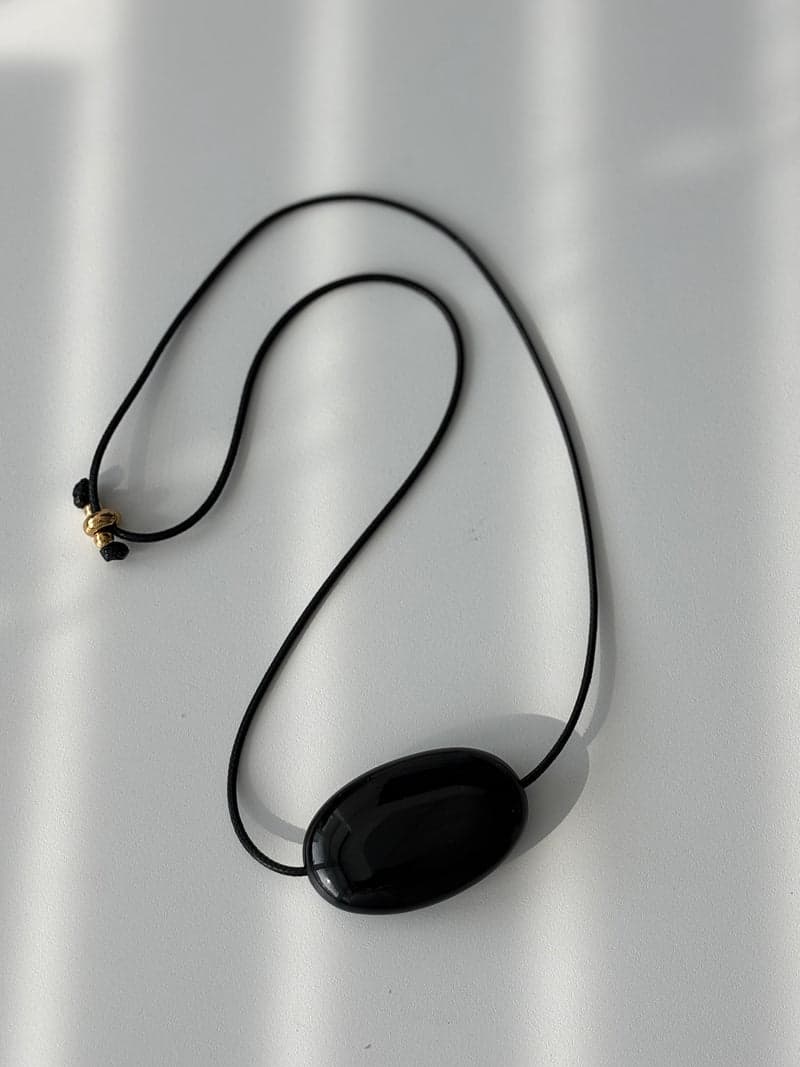 A single large flat glossy black oval pendant on a thin black cord, lying on a white surface in window light