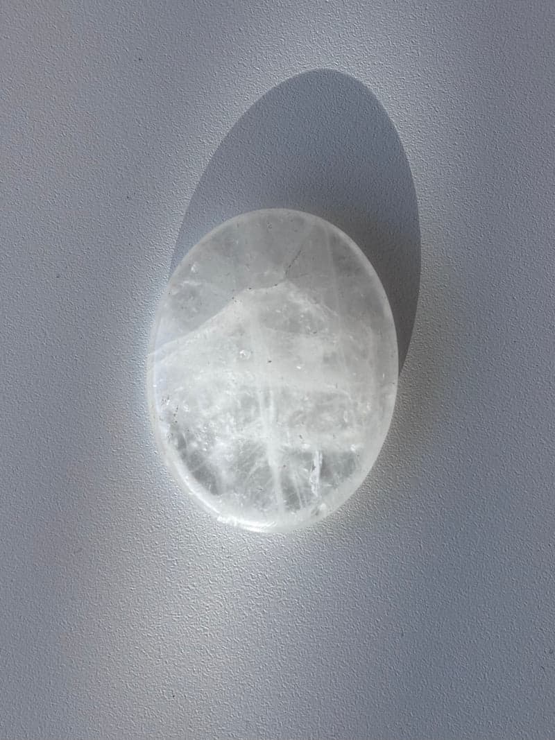 Ein poliertes Oval aus klarem Quarz mit weißen inneren Schleiern, auf weißem Untergrund liegend und einen weichen Schatten werfend