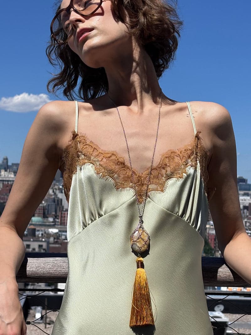 A woman on a rooftop in a pale green slip dress wearing the long mesh necklace with a warm toned stone and a gold tassel