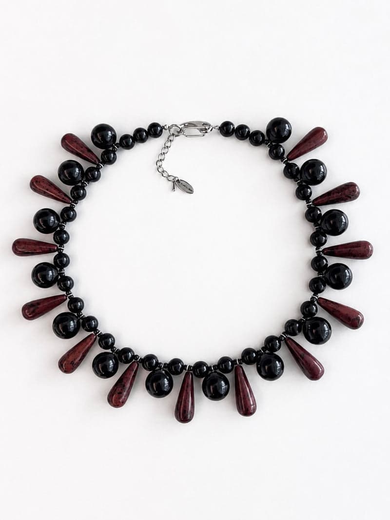 Um colar peitoral de contas pretas redondas e brilhantes alternadas com longas contas vermelho-escuras em gota, sobre um fundo branco