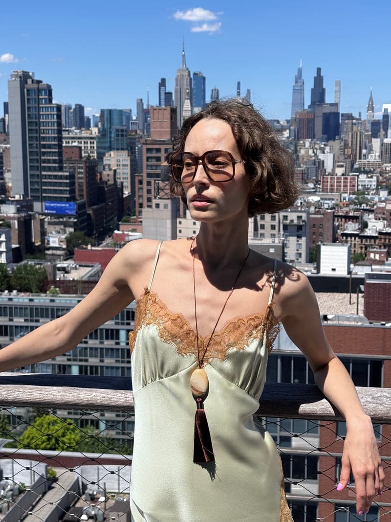 Uma mulher em um terraço na cidade, em um vestido slip verde-claro, usando um cordão longo com uma pedra oval bege e uma borla marrom