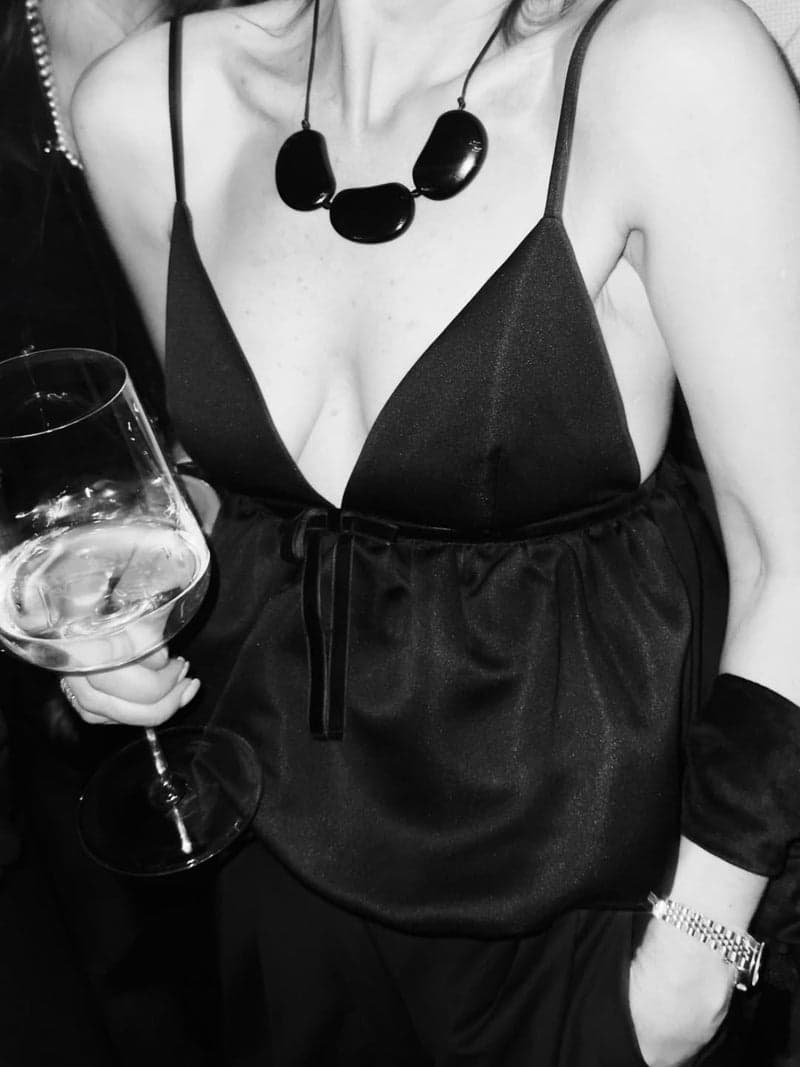 Uma fotografia em preto e branco de uma mulher em um top slip preto segurando uma taça de vinho, usando um colar de três grandes pedras ovais escuras