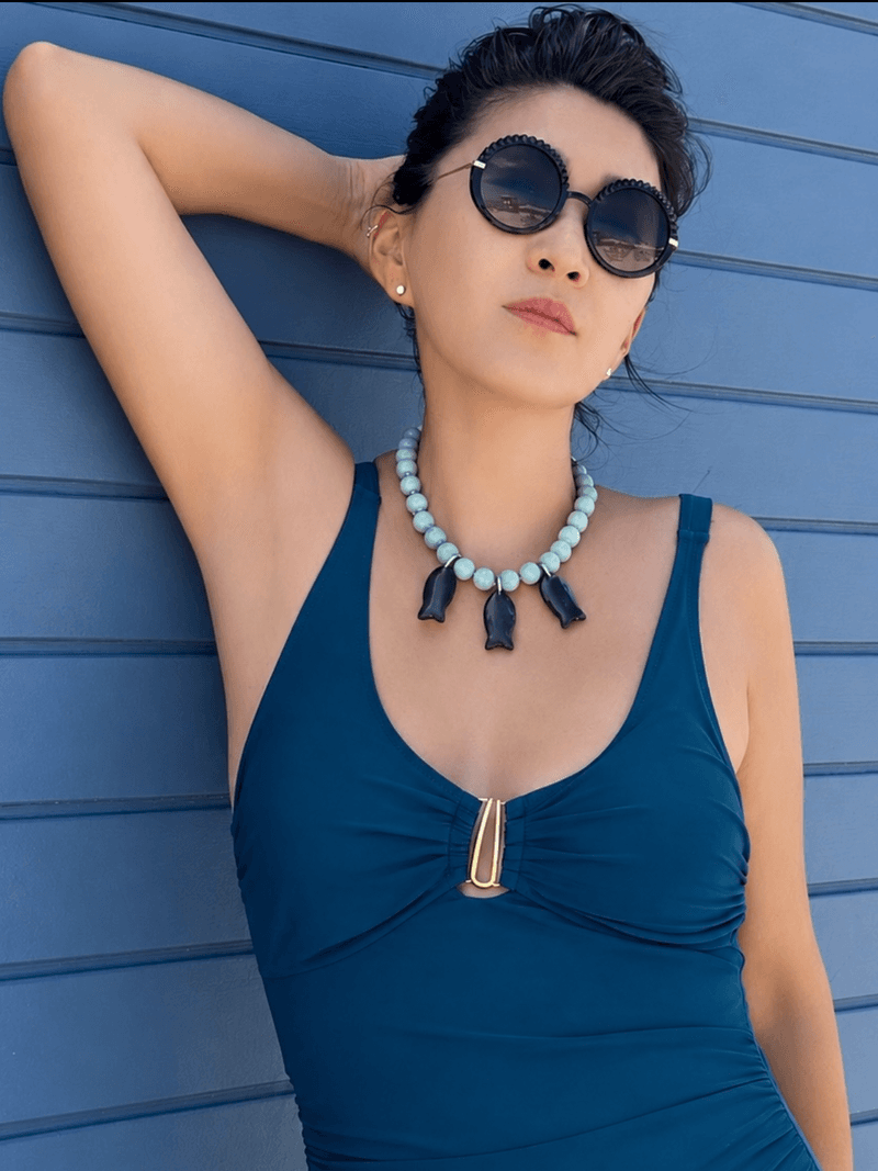 Eine Frau mit Sonnenbrille und petrolfarbenem Badeanzug vor blauer Wand, mit einer blassgrünen Perlenkette mit drei dunklen Fisch-Anhängern