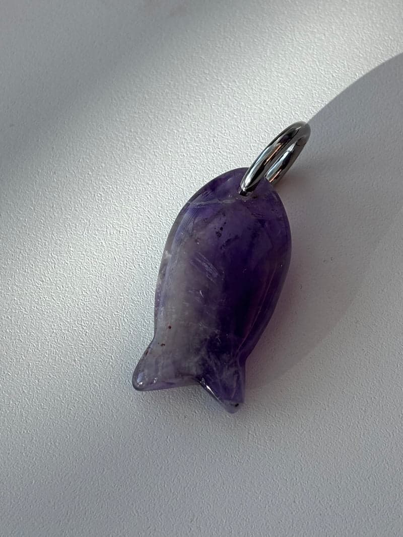 Ein kleiner geschnitzter Fisch-Anhänger aus durchscheinendem violettem Stein mit einem silbernen Ring, auf weißem Untergrund im Fensterlicht