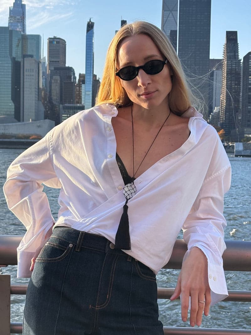Eine Frau mit Sonnenbrille und offenem weißem Hemd an einer städtischen Uferpromenade, mit der langen Kordelkette mit weißem Quarzstein und schwarzer Quaste
