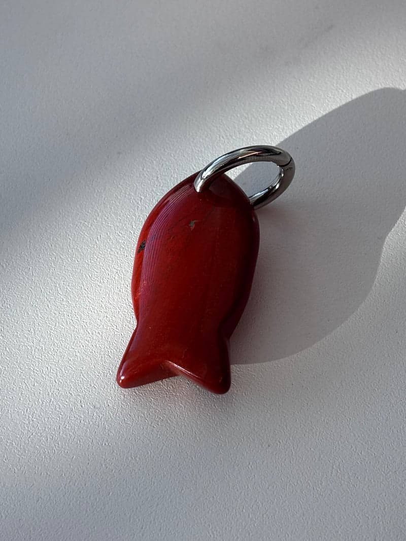 Um pequeno pingente de peixe entalhado em pedra vermelho-escura brilhante com um anel prateado, sobre uma superfície branca sob luz de janela
