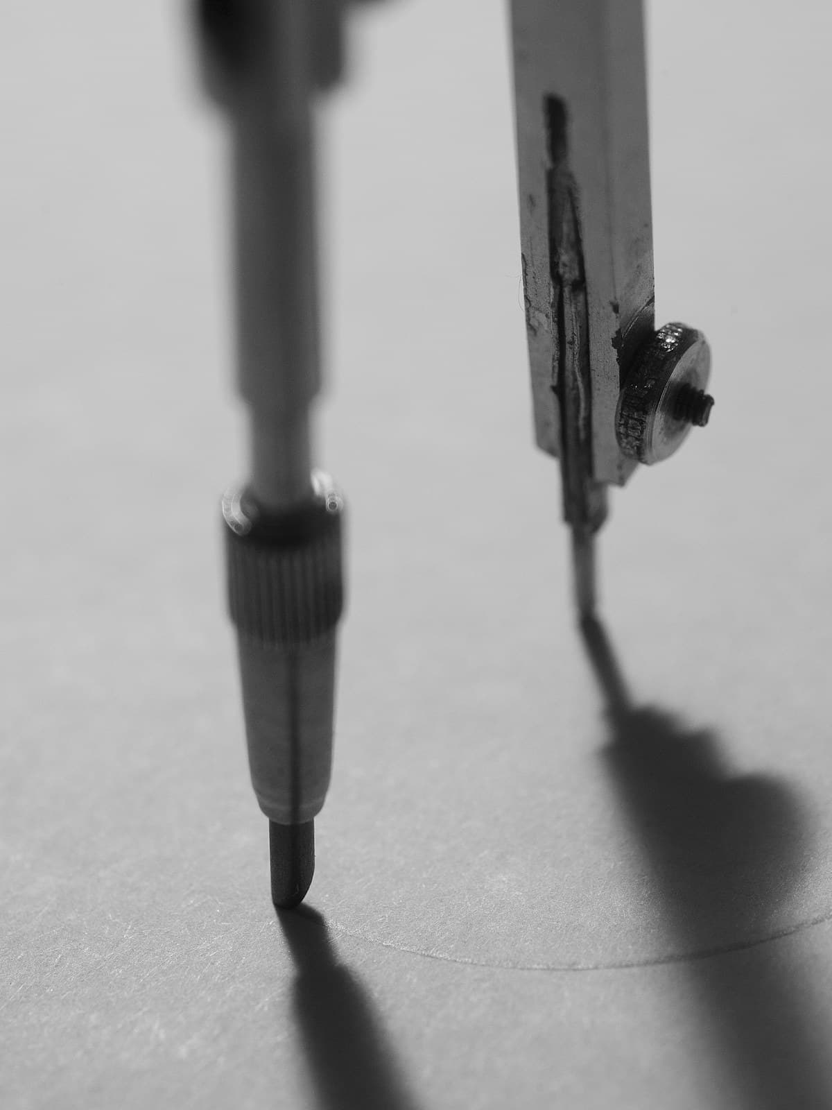 A metal drawing compass drawing a circle on white paper, in black and white