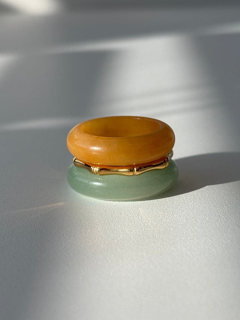 A stacked ring set in sunlight: an orange stone band on top, a thin gold bamboo-shaped ring, and a pale green stone band below