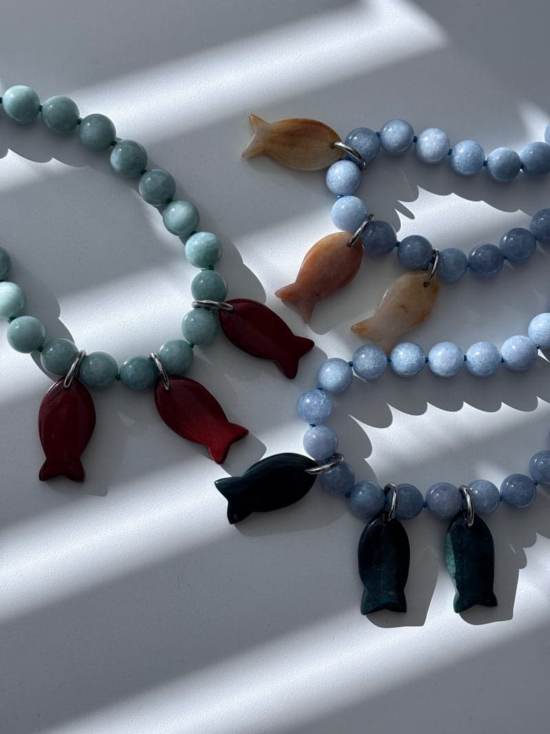 Several bead necklaces in blue and green laid together, each with small carved stone fish pendants in different colours