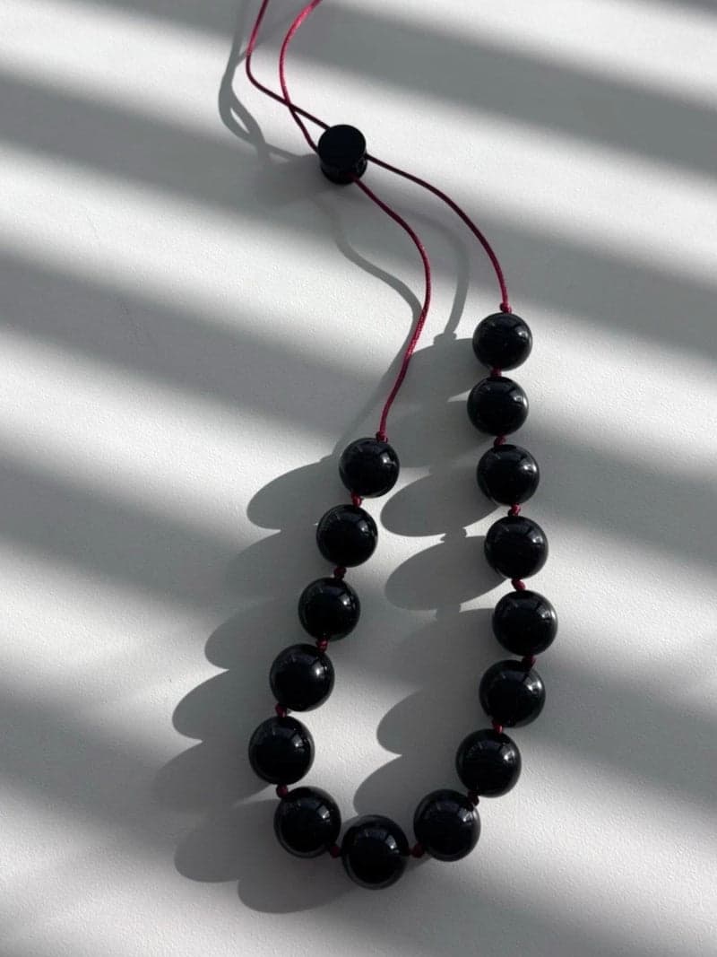 Um colar de grandes contas redondas pretas e brilhantes enfiadas em um fino cordão vermelho, disposto em laço sobre uma superfície branca sob luz listrada de janela