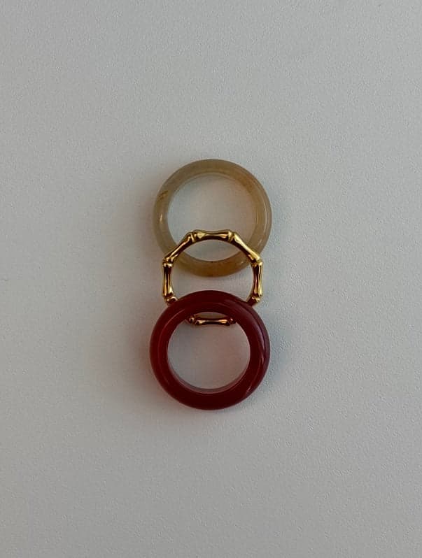 Deux bagues en pierre et un fin anneau doré en forme de bambou disposés en ligne verticale : un anneau jaune pâle, l'anneau doré, et un anneau rouge profond