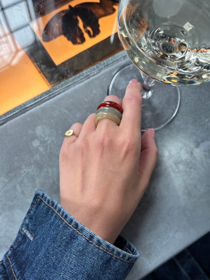 A hand resting beside a wine glass on a grey table, wearing the red, green and yellow stone rings stacked on one finger