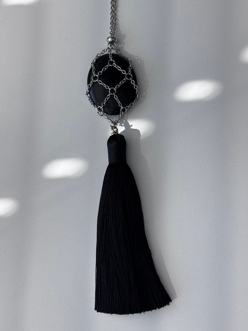 Ein ovales schwarzes Obsidian-Stück, gehalten in einem silbernen Kettengeflecht über einer schwarzen Quaste, vor einer weißen Wand hängend