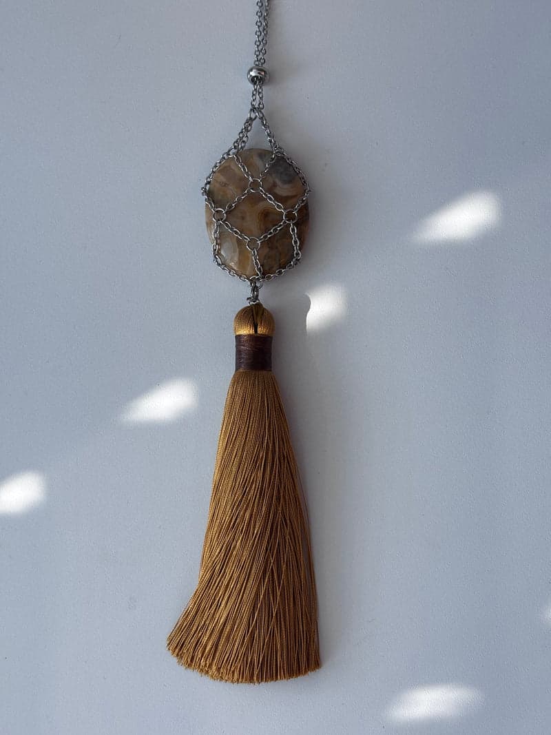 Une agate ovale rubanée crème, beige et grise tenue dans une résille de chaîne argentée au-dessus d'un long pompon brun doré