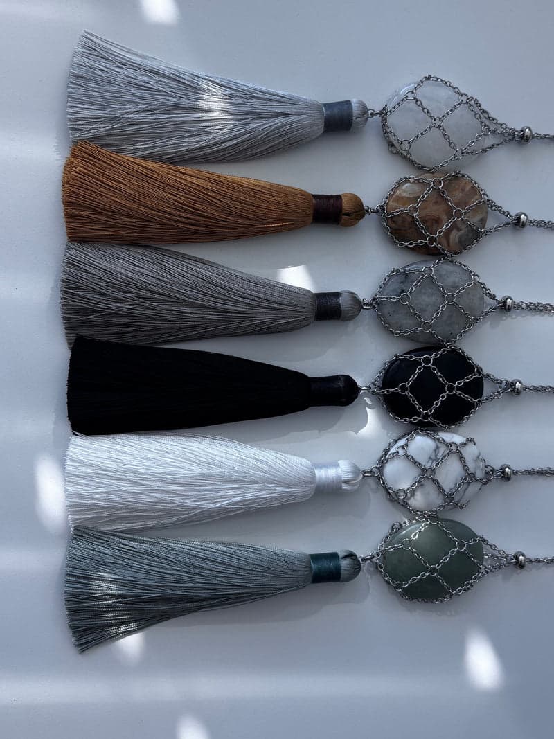 Six silver chain mesh pendants laid side by side with tassels, each holding a different oval stone: clear, tan, grey, black, white and green